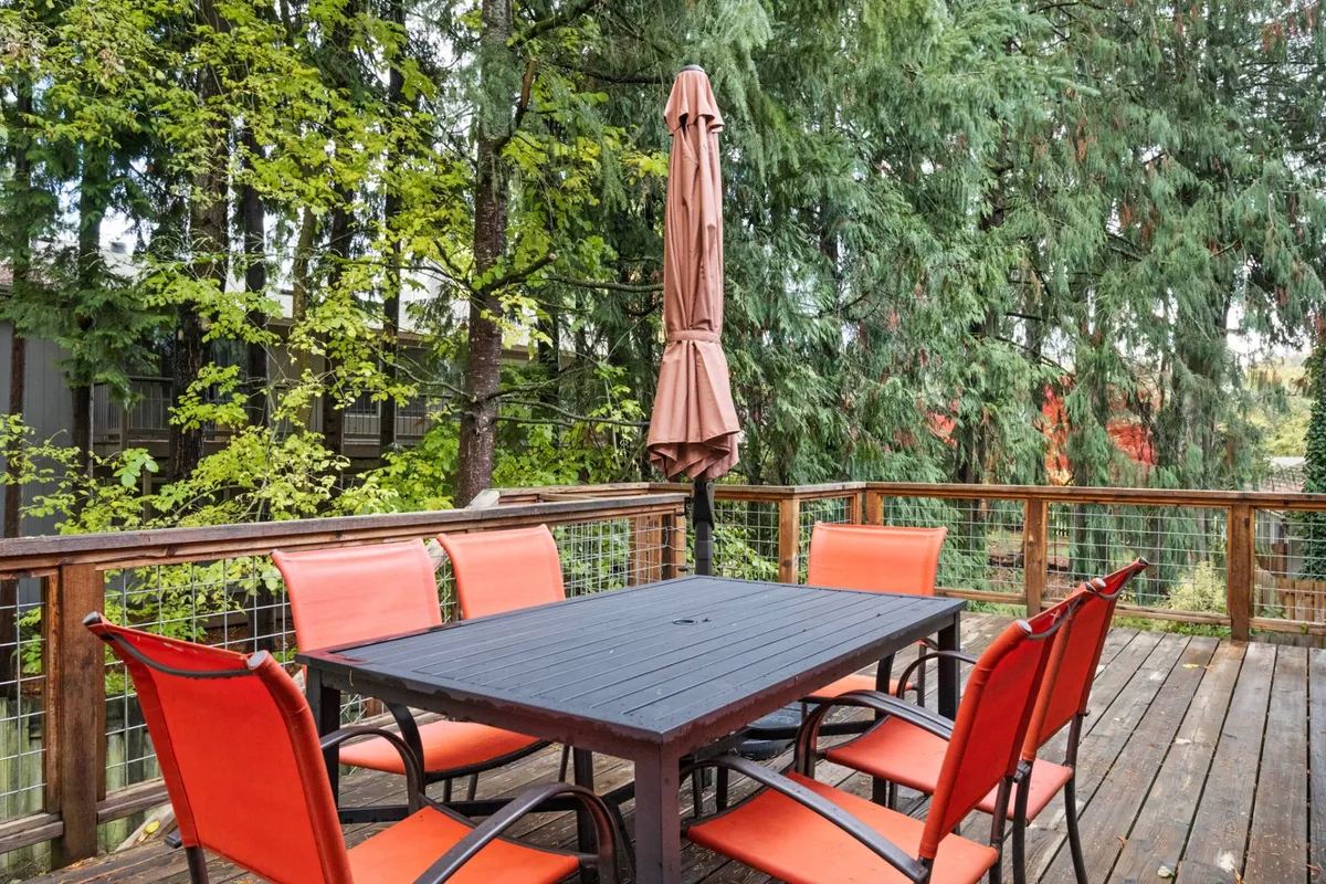 An exterior shot of a spacious wooden deck at Bridlemile Cottage, featuring a wooden table, bright red chairs, and a large parasol. The area is surrounded by lush greenery, creating a casual and inviting outdoor setting perfect for relaxing with pets or enjoying meals al fresco.