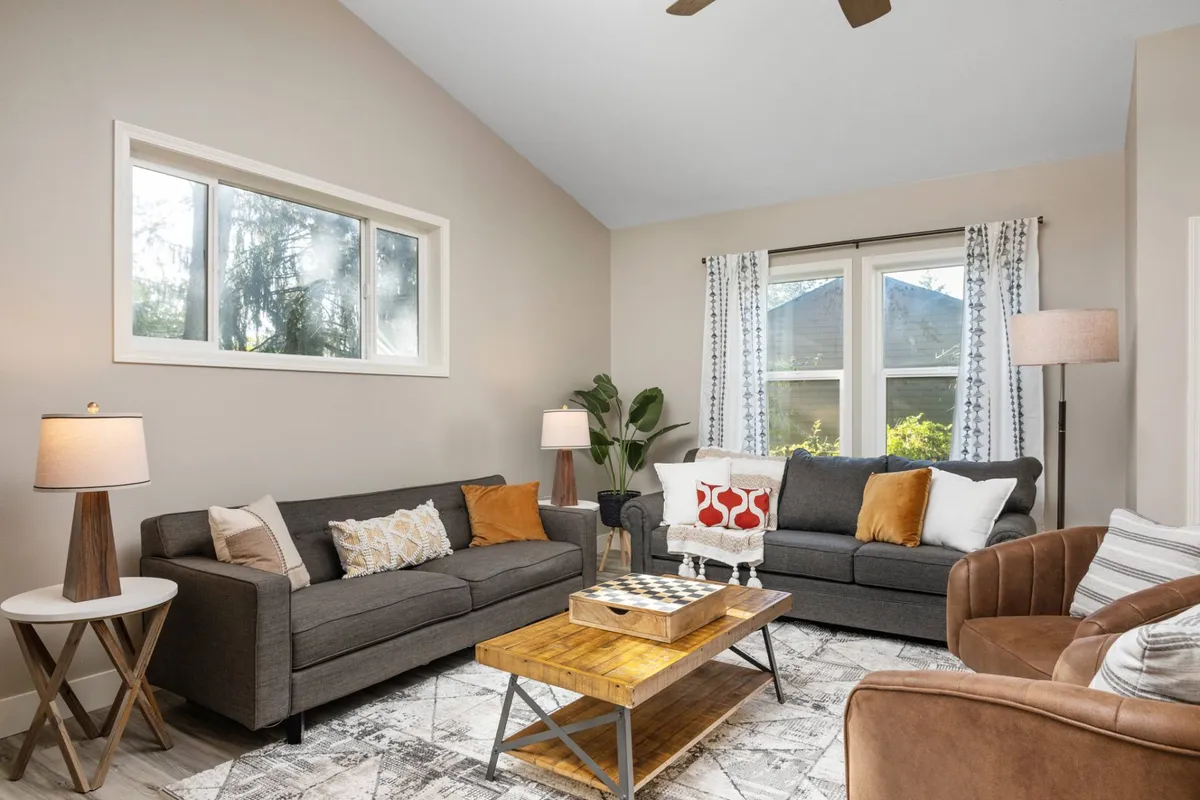 A spacious living area adorned with inviting sofas, a central coffee table, and an assortment of lamps. The windows allow ample natural light to pour in, illuminating lush plants and creating a lively yet relaxing atmosphere ideal for group activities and family time.