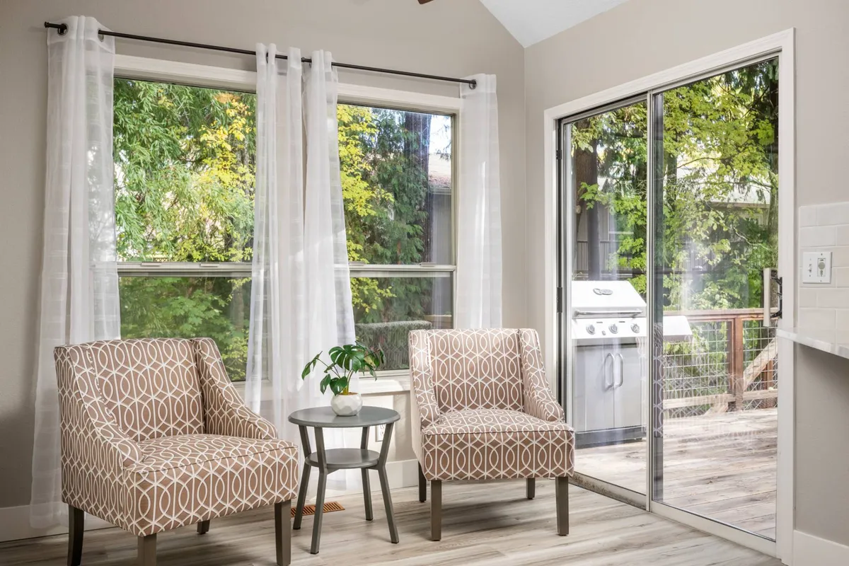 A cozy sitting nook with two patterned chairs and a small round table is shown by large windows, sheer curtains, and a sliding door leading to the deck. The space is bright and ideal for relaxing, having coffee, or planning the day.