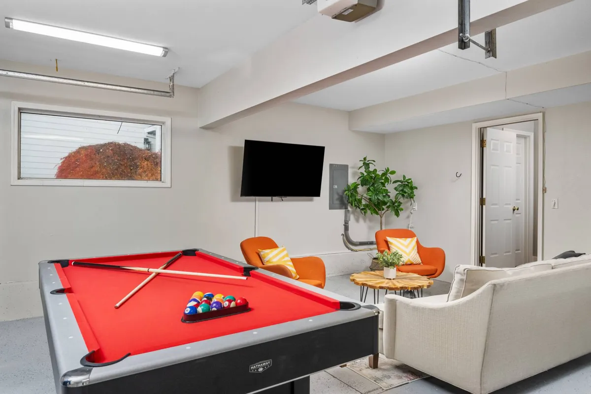 This image captures the heart of the converted garage's entertainment space, featuring a pool table at the center, bold orange chairs providing comfortable seating, a large television for streaming, and a green plant that adds a touch of vibrancy. The layout suggests a lively area perfect for group activities, games, and relaxing together.