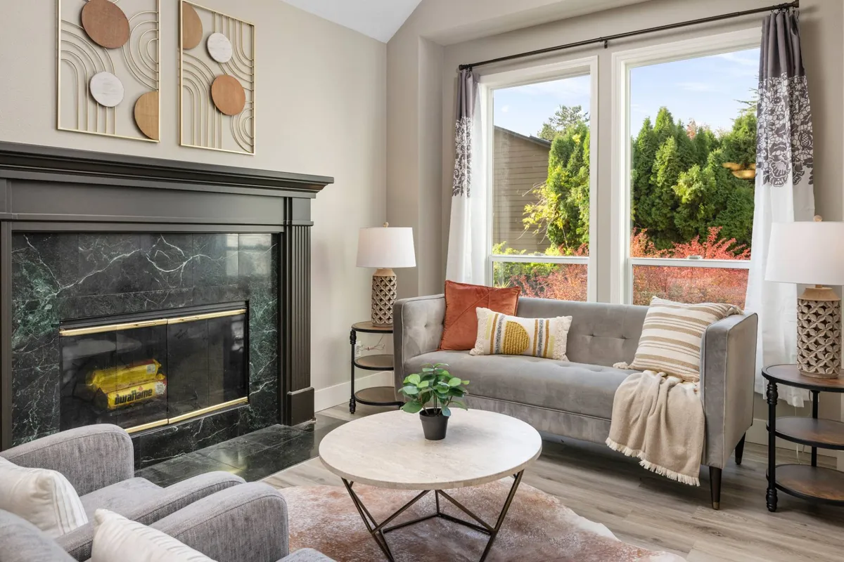 A warm, inviting living area featuring a plush sofa and coffee table set before a glowing fireplace. Large windows fill the room with natural light, offering tranquil views of greenery outside. The minimalist decor emphasizes comfort and togetherness, with cozy seating arranged to encourage conversation or relaxation.