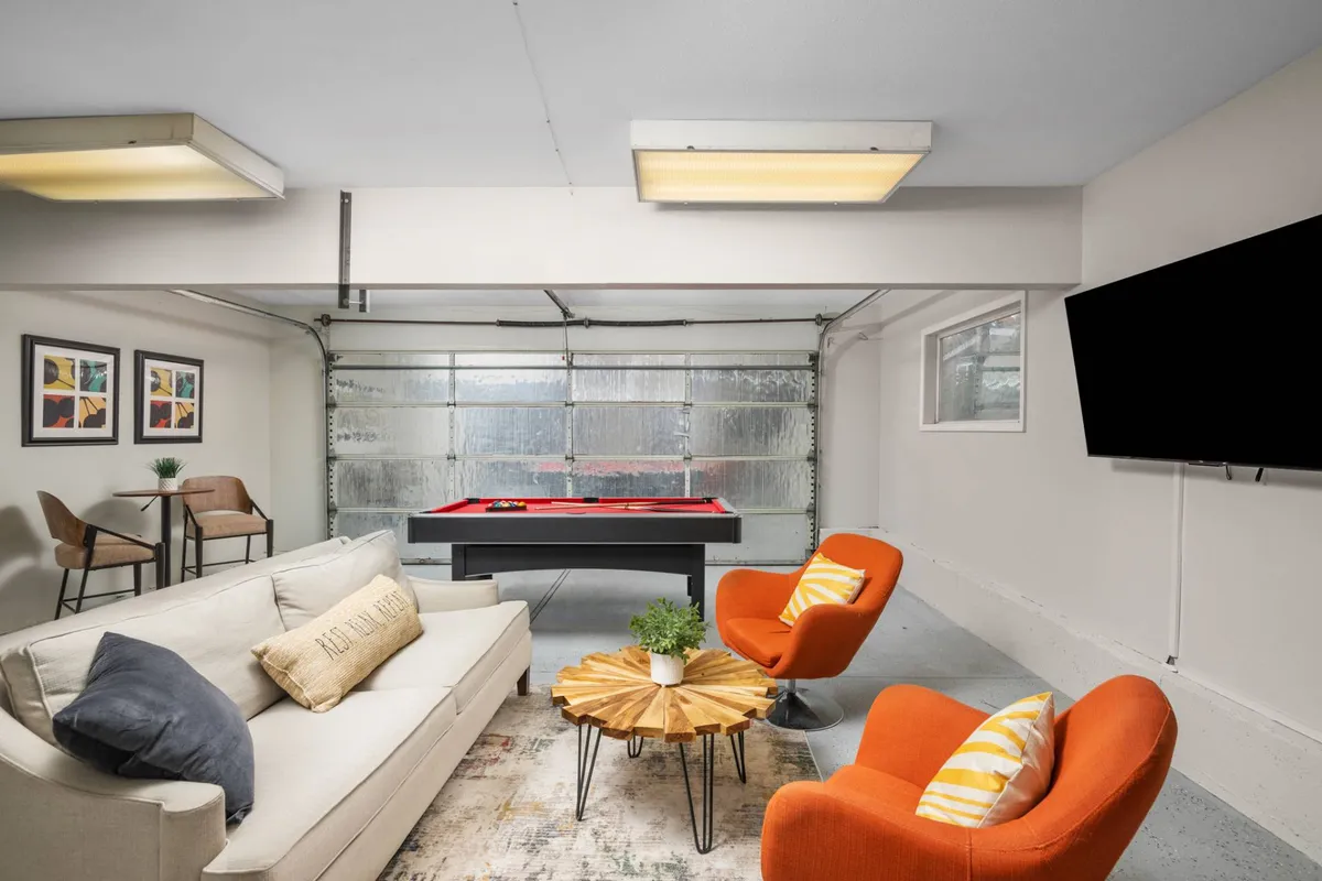 A broader view of the converted garage reveals the pool table from another angle, cozy orange chairs, a plush sofa, and wall art that enhances the space's playful vibe. The overall setup encourages both active play and relaxed lounging.