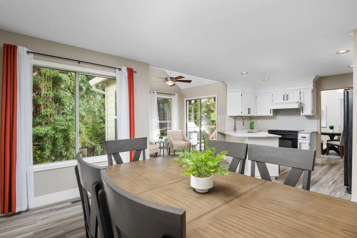 An open-plan dining space is pictured, with a dining table set near large windows that allow ample sunlight to pour in. The kitchen area is visible in the background and the outdoor view adds a natural, vibrant backdrop.