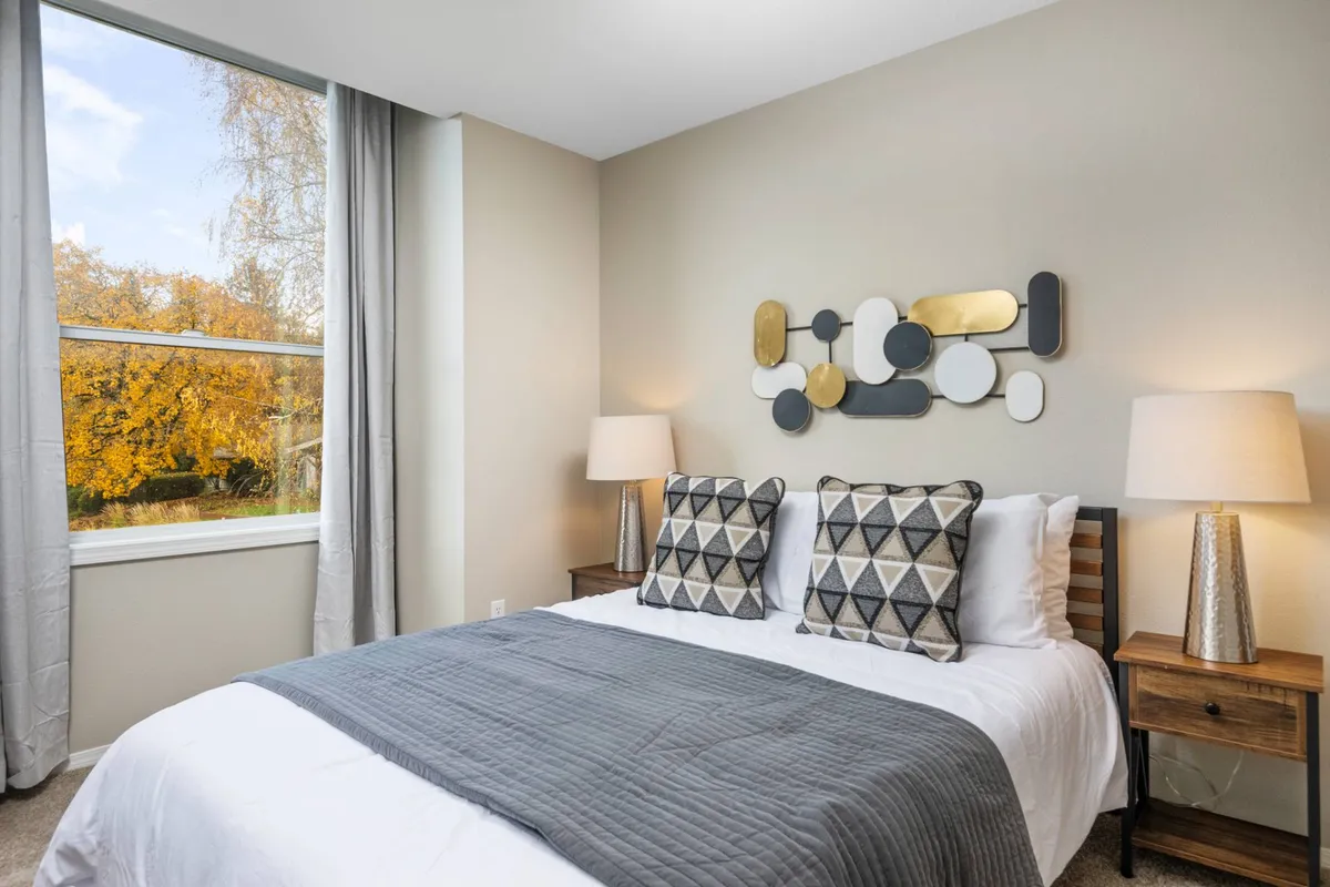 A bedroom flooded with natural light from a large window, featuring a neatly made bed with decorative pillows and abstract wall art. The room feels airy, peaceful, and ideal for relaxation or reading by the window.