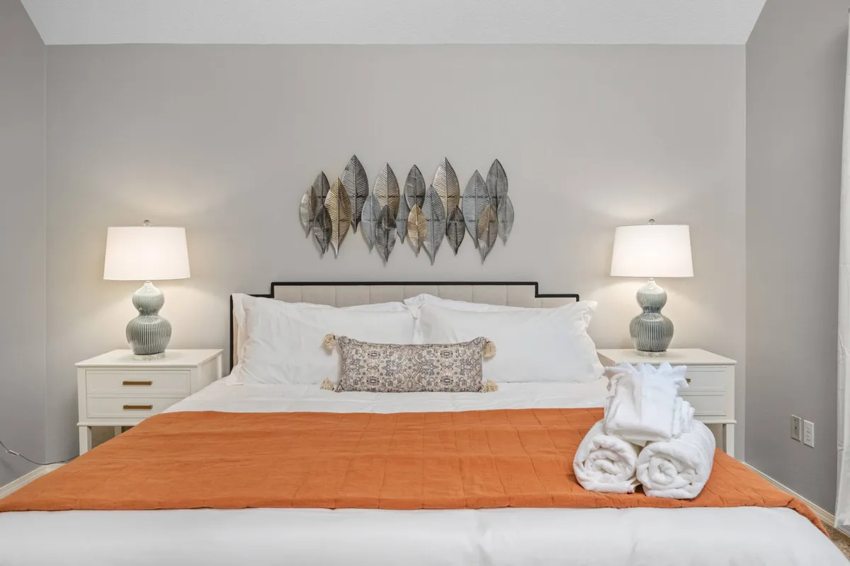 A cozy bedroom setting featuring a bed with plush pillows, decorative wall art above the headboard, and matching nightstands with table lamps on either side. The space is softly lit, offering a welcoming and restful ambiance ideal for unwinding at the end of the day.