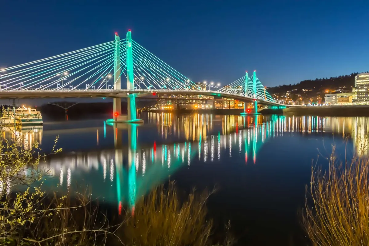 A stunning exterior shot featuring one of Portland's iconic bridges aglow with city lights, reflecting beautifully across the river. The image captures the energy of the city as evening falls, with a tranquil water foreground and a lively urban skyline.