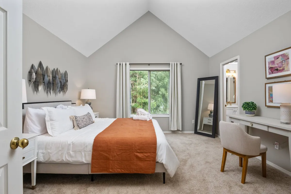 A spacious bedroom with a bed covered in a soft orange throw blanket, positioned beside a large window overlooking lush greenery. Decorative wall art adds a personal touch, while a desk and chair create a nook for relaxing or working. Natural light fills the room, enhancing its tranquil and inviting atmosphere.