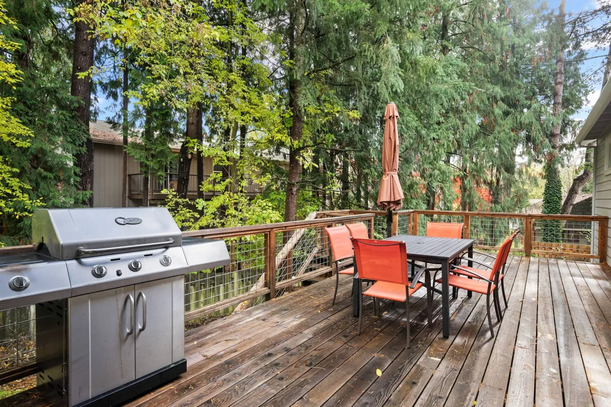 A welcoming backyard deck setup featuring a grill, dining table, chairs, an umbrella for shade, and lush surrounding trees. The space suggests a casual, family-friendly atmosphere ideal for outdoor meals, play, and relaxation after a day of outings.